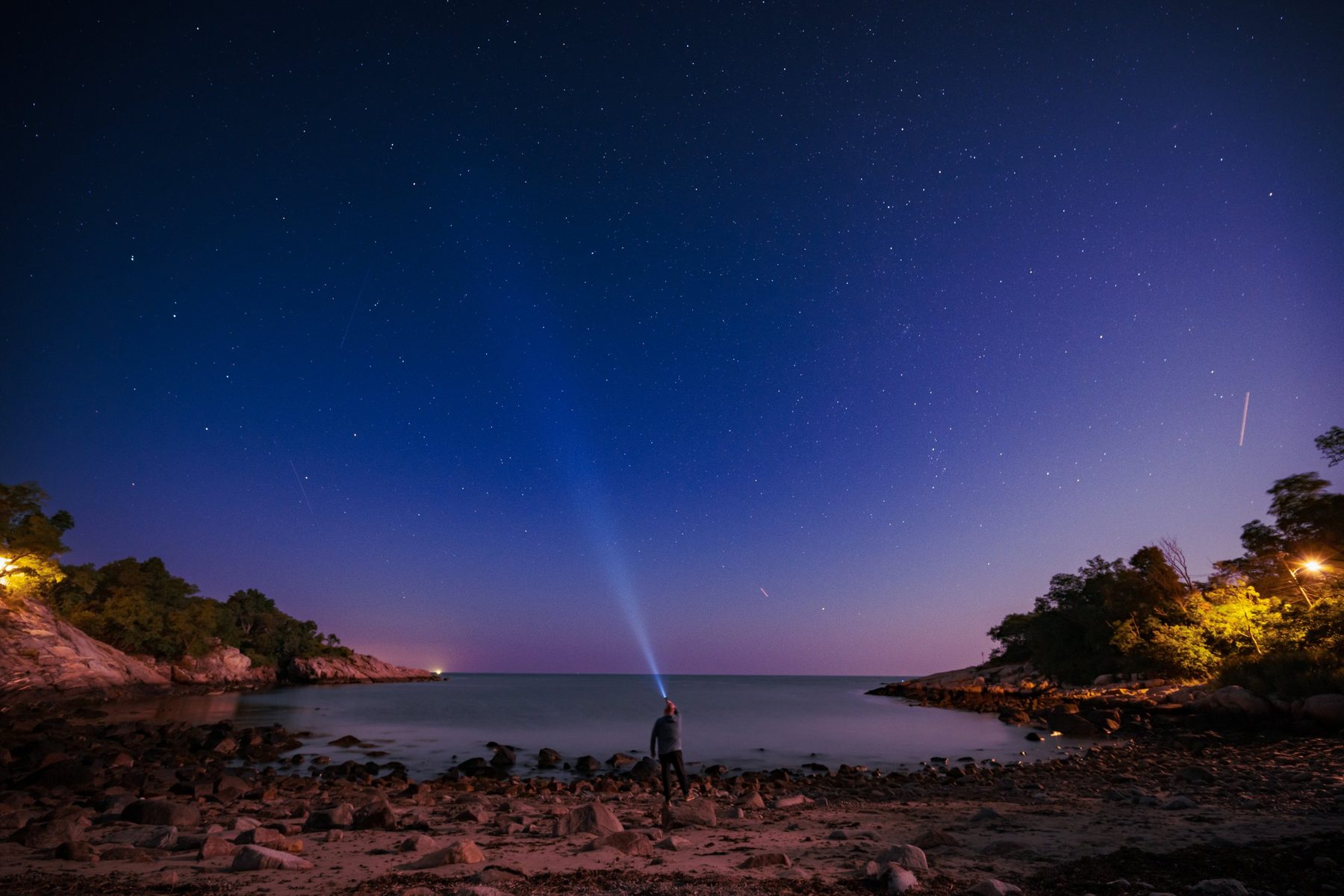 Jerrame standing on a rocky New Hampshire beach at night, shining a light up into a sky full of stars