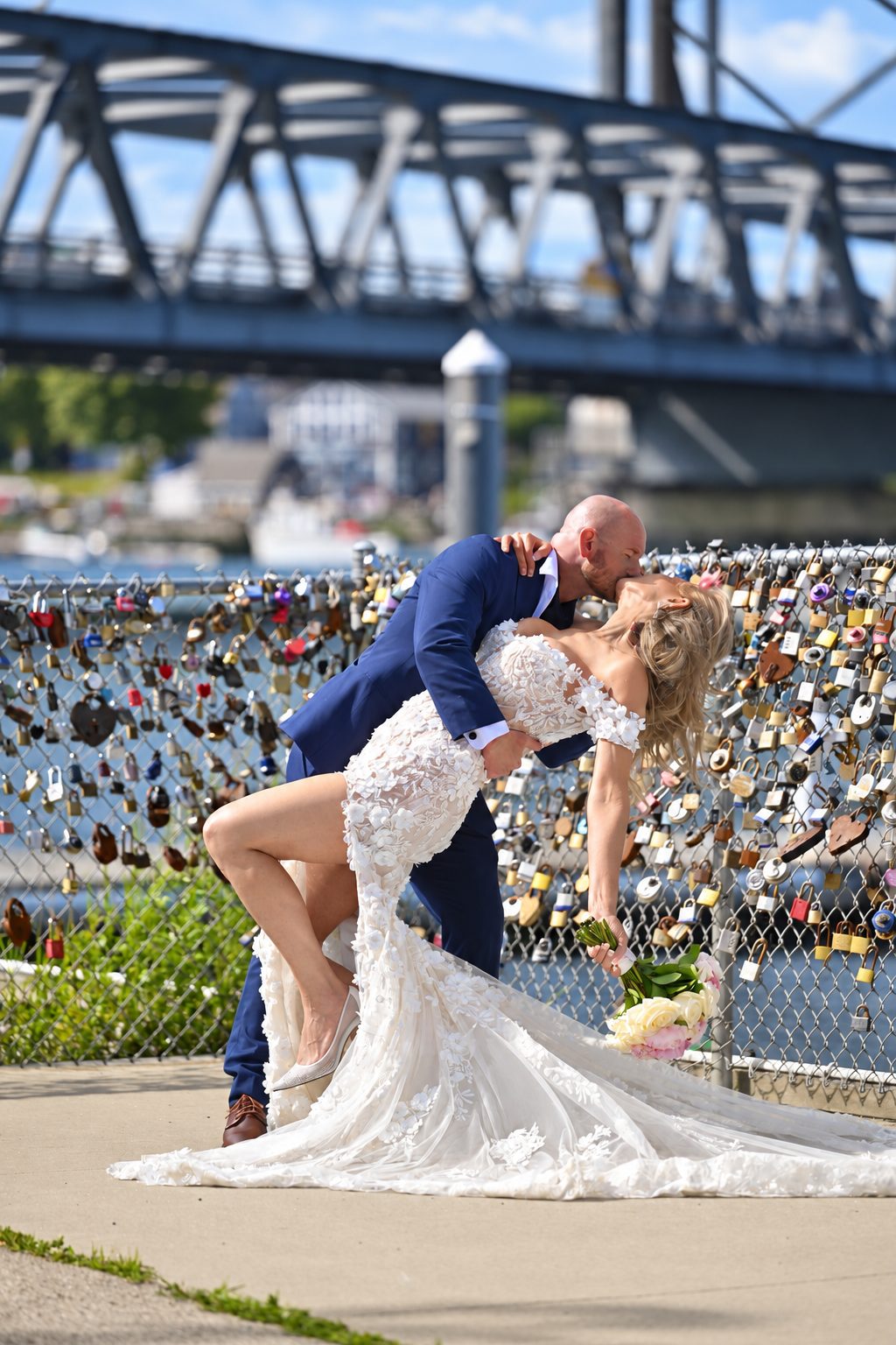 Bride and groom dip and kiss beside a bridge covered in locks